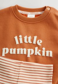 Rust orange baby sweater with white text "little pumpkin" and matching cream and orange striped pants. Small button detail on shoulder.