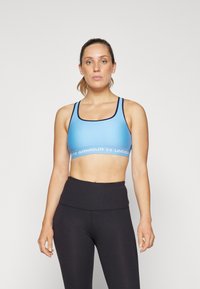 Lyseblå sports-bh med scoop-hals, marineblå kant og hvit "UNDER ARMOUR" logo-bånd. Kombinert med høyt liv sorte tights.