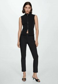 Gilet nero senza maniche, aderente, con colletto alto e zip frontale, abbinato a pantaloni neri sartoriali e sandali con tacco aperti neri.