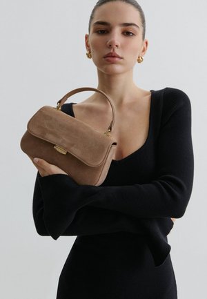 Jeune femme en robe noire à encolure dégagée, tenant un sac à main en daim marron avec une anse supérieure et une fermeture dorée, sur un fond uni.