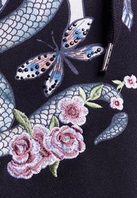 Tela negra con intrincados bordados de rosas en tonos de rosa y púrpura, una mariposa multicolor y escamas en tonos de azul y verde.