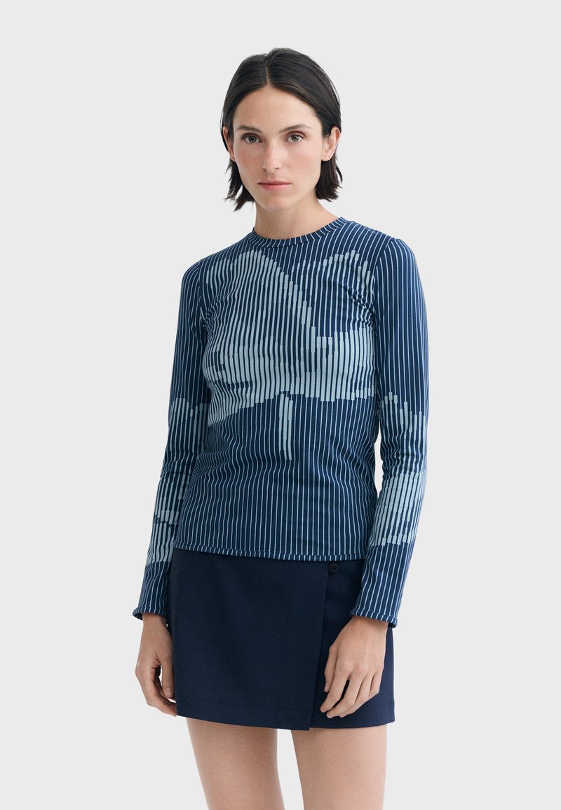 Femme aux cheveux courts et foncés portant un haut à manches longues rayé bleu et blanc ainsi qu'une jupe bleu marine, debout devant un fond uni.