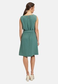 Grünes ärmelloses Kleid mit einem Taillengürtel, strukturiertem Stoff und einem kleinen Ausschnitt hinten. Der Saum reicht bis über das Knie.