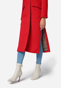 Manteau long rouge au design structuré, avec une doublure en imprimé léopard. Assorti avec un jean skinny bleu clair et des bottines beiges.