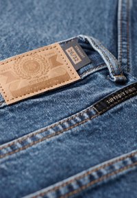 Jeans de mezclilla que presentan una etiqueta de cuero marrón claro marcada con "Superdry" y detalles en relieve, etiqueta de tamaño que dice "26/32" y costuras visibles en un hilo oscuro.