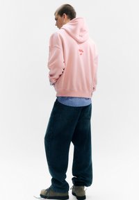 Rózsaszín kapucnis pulóver piros logóval a hátán, alatta kék csíkos ing. Bő fazonú sötét farmernadrág és szürke cipő egészíti ki az öltözéket.