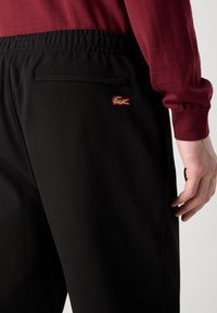 Zwarte textuurbroek met een achterzak en een klein, rood en gouden geborduurd krokodillenlogo aan de rechterzijde.