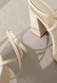 Cremefarbene High-Heelsandalen mit einem strukturierten Krokodilmuster, quadratischem Blockabsatz und einem zierlichen Knöchelriemen mit einer kleinen goldenen Schnalle.