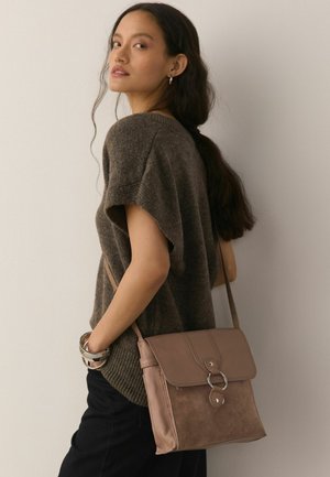 Kvinde med langt mørkt hår iført en brun strikket top, sorte bukser, sølvarmbånd og en lysebrun læder- og ruskindskrossbodytaske.