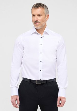Weißes Langarmhemd mit schmalem Kragen, versehen mit schwarzen Knöpfen auf der Vorderseite. Getragen mit schwarzen Hosen und einem passenden Gürtel.