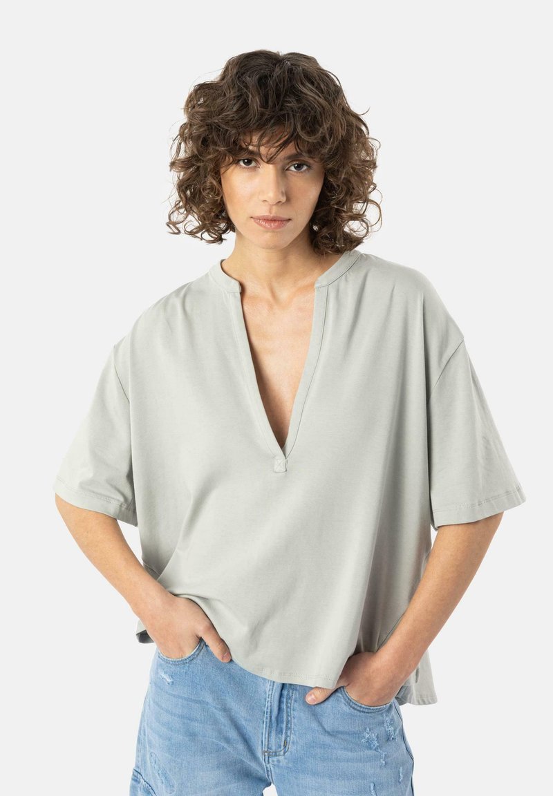 Chemise gris clair à manches courtes avec un col en V et un accent bouton. Texture douce et lisse, coupe décontractée et design légèrement court.