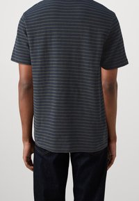 Personne portant une chemise à manches courtes bleu marine à fines rayures horizontales et un jean bleu marine, vue de dos sur un fond uni.