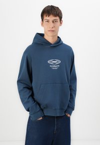 Jaunas vyras, vilkintis mėlyną džemperį su "1990 CoreSport Athletic Essentials Creative Dept" logotipu ir tamsiai mėlynomis džinsomis, rankos kišenėse, neutralus fonas.