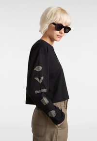 Zwarte katoenen cropped langesleevenshirt met zilveren graphics op de mouwen, ronde halslijn en een relaxte pasvorm. Zonnebril maakt de look compleet.