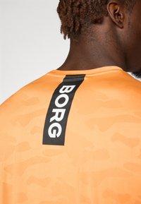 Orange atletisk skjorte med et struktureret, camouflagemønster; har et sort lodret logobånd mærket "BORG" på ryggen.