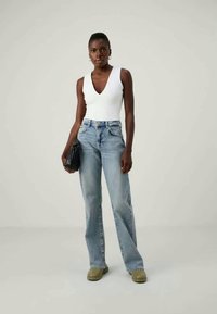 Bijela bezrukavna majica, svijetloplave traperice visoko strukirane, tamnozelene cipele bez pertli. Model dr�ži crnu torbu, stoji naslonjen/a na neutralnu pozadinu.