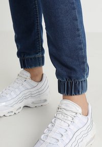 Pantalones jogger de mezclilla azul con puños elásticos, combinados con zapatillas blancas que presentan detalles transparentes y un diseño a rayas en los costados.