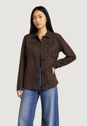 Donna con indosso una giacca marrone scuro con frange, jeans blu a gamba larga, con una mano in tasca su uno sfondo chiaro uniforme.