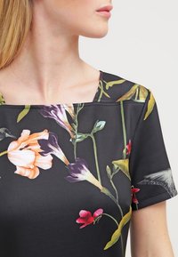 Blouse à manches courtes avec un décolleté carré, fabriquée en tissu noir ornée de motifs floraux vibrants dans des teintes de rose, orange, violet et vert.