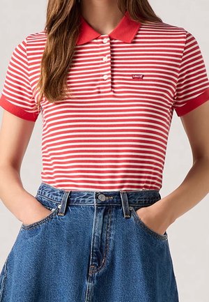 Kvinde iført rød- og hvidstribet polotrøje med rød krave og blå højhåndede jeans med hænderne i lommerne.