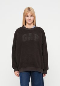 Sudadera de forro polar marrón oscuro con superficie texturizada, con un logo "GAP" en relieve en el frente, puños y dobladillo acanalados.