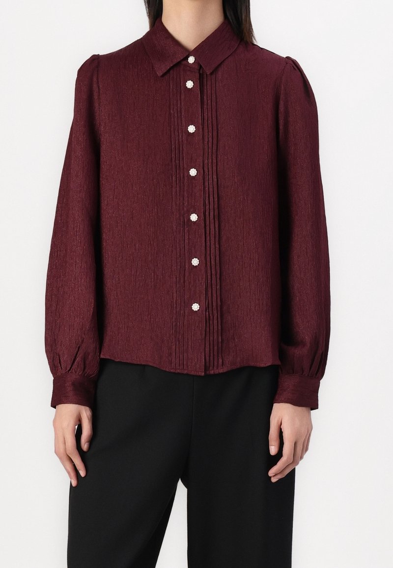 Blouse bordeaux avec une finition texturée, devant boutonné avec des accents en perles, et manches longues bouffantes ; design plissé le long de la patte de boutonnage.