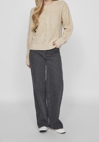 Beige kabelstickad tröja med rund halsringning, kombinerad med vida mörkgrå jeans. Tröjan har texturerade mönster och ribbade ärmslut.