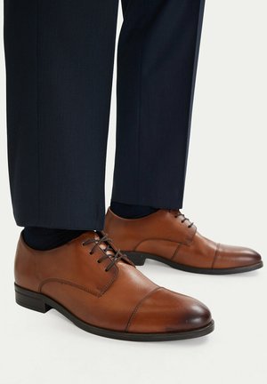 Zapatos de vestir para hombres de cuero marrón con cordones, usados con pantalones azul oscuro y calcetines negros sobre un fondo claro.