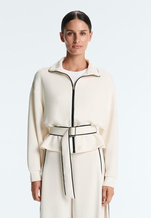 Vrouw draagt een crèmekleurig jack met ritssluiting en zwarte bies, met bijpassende broek met ceintuur, staand tegen een egale lichte achtergrond.