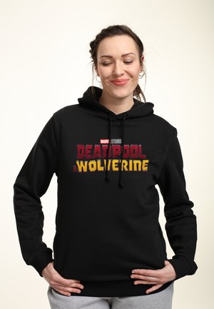 Kvinde med fletninger, iført en sort Marvel Studios Deadpool og Wolverine hættetrøje, smilende med hænderne hvilende på hofterne.
