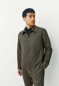 Giacca marrone con zip realizzata in tessuto testurizzato, caratterizzata da un colletto a punta e tasche laterali, abbinata a una camicia button-up scura.