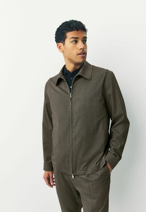 Chaqueta marrón con cremallera, hecha de tejido texturizado, con cuello puntiagudo y bolsillos laterales, combinada con una camisa de botones oscura.