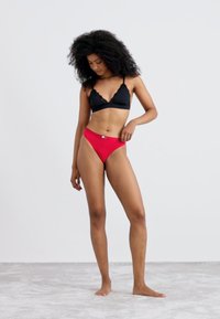 Musta pitsireunainen bralette ja punaiset bikinit, jotka tarjoavat sileän tekstuurin ja istuvan muotoilun, päällä mallilla neutraalilla taustalla.