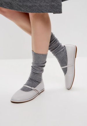 Weiße Ballerinas aus Leder mit einer runden Zehenpartie, ausgestattet mit einem dünnen Riemen und einer hellbraunen Sohle. Kombiniert mit gerippten grauen Kniestrümpfen.