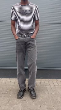 Uomo che indossa una t-shirt Guess Jeans USA a righe bianche e nere, jeans neri sbiaditi, mocassini neri, in piedi con le mani in tasca contro un muro grigio.
