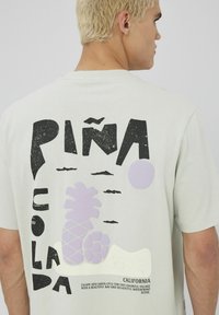 T-shirt gris clair avec un graphisme "Piña Colada" en noir vif et lavande douce, accompagné d'une illustration d'ananas et de détails textuels.