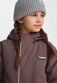 Braune wasserdichte Jacke mit Kapuze, versehen mit einem weißen Reißverschluss und dem "hummel"-Logo. Kombiniert mit einer gerippten grauen Mütze. Lange geflochtene Haare.