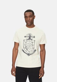 Camiseta de hombre de color crema hecha de algodón, con un diseño gráfico que incluye un escudo, un ancla y el texto "Desde Guess 1981" en tinta negra.
