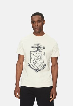 Camiseta de hombre de color crema hecha de algodón, con un diseño gráfico que incluye un escudo, un ancla y el texto "Desde Guess 1981" en tinta negra.