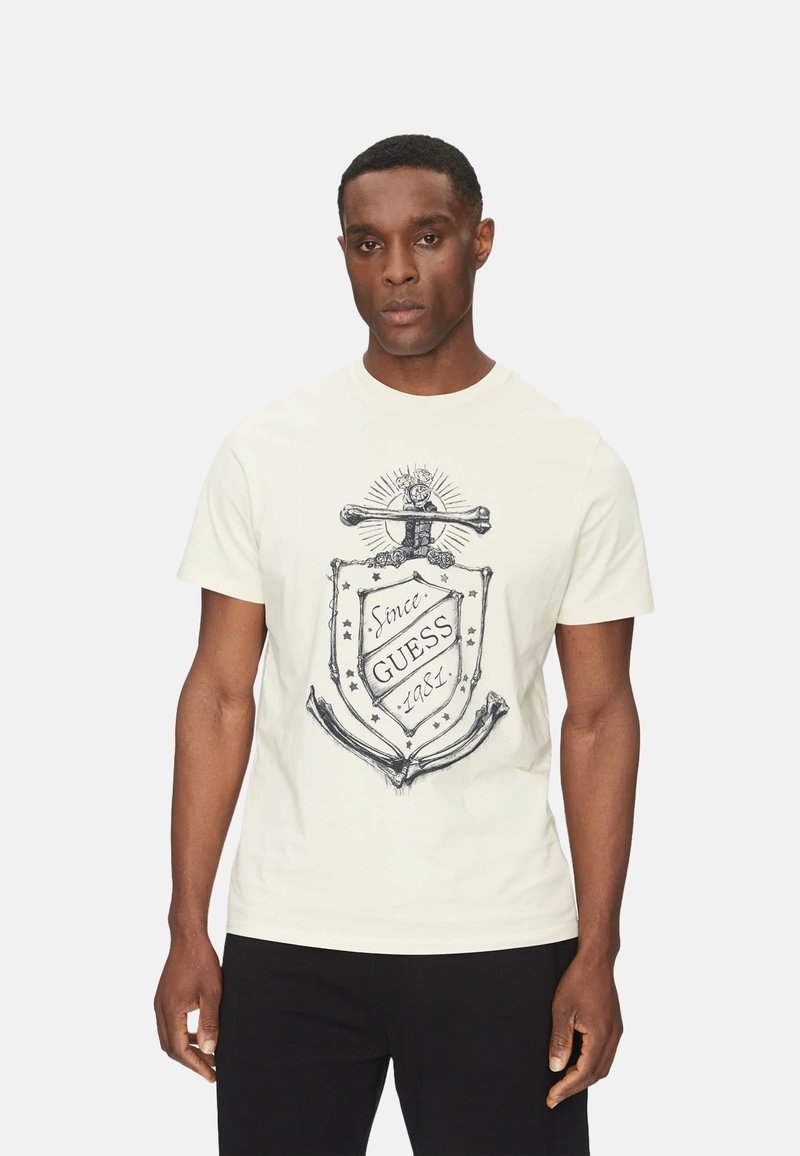 Camiseta de hombre de color crema hecha de algodón, con un diseño gráfico que incluye un escudo, un ancla y el texto "Desde Guess 1981" en tinta negra.