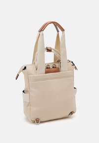 Bolsa tote de lona beige con asas de tela anchas, ribete de cuero, anillos de metal, bolsillo frontal y cierre de cremallera, mostrada sobre un fondo blanco.