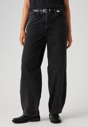 Persoon die losse zwarte denimspijkerbroek, zwarte riem, zwart topje en zwarte enkellaarsjes draagt, staand tegen een effen lichte achtergrond.