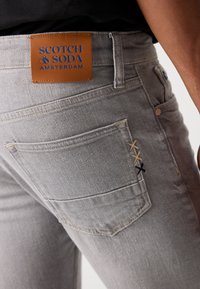 Grijze denim jeans met een leren label met de merknaam "SCOTCH & SODA AMSTERDAM" en decoratieve stiksels op de achterzak.