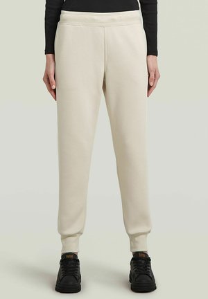 Beige sweatpants with a ribbed waistband, tapered legs, and elastic cuffs, paired with black sneakers. Smooth texture, casual design.