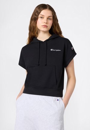 Mujer joven con cabello largo que lleva una sudadera negra de manga corta de Champion y pantalones deportivos grises claros de Champion, con las manos en los bolsillos.