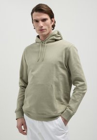 Uomo che indossa una felpa con cappuccio verde chiaro con tasca frontale e pantaloni bianchi, in piedi su uno sfondo chiaro e semplice.