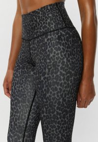 Detailný záber osoby, ktorá má na sebe vysoké čierno-sivé legíny s leopardím vzorom a malým okrúhlym logom blízko pásu.