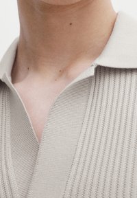 Polo en maille beige présentant une texture côtelée et un col décontracté. La vue en gros plan met en évidence le matériau doux et les détails subtils.