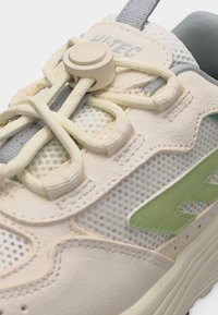 Bež tenisica od mrežaste tkanine, sa svijetlozelenim bočnim logotipom, bež podesivim kopčom za vezice i debelim potplatom, prikaz izbliza.