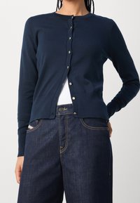Tmav�ě modrý pletený kardigan s kulatým výstřihem, zapínáním na knoflíky a žebrovanými manžetami. V kombinaci s tmavými džínami z denimu s kontrastním prošíváním.